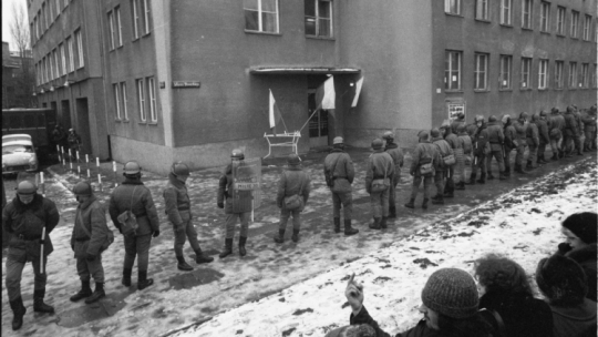 Warszawa, 1981-12-02. Rozpoczynają się przygotowania do zdławienia strajku studentów Wyższej Oficerskiej Szkoły Pożarnictwa w Warszawie przy ul. Słowackiego 52/54, którzy domagali się nadania cywilnego charakteru uczelni. Nz. zomowcy kordonem otaczają budynek WOSP. Mężczyzna obserwujący sytuację wykonuje w ich stronę nieprzyzwoity gest. Fot. PAP/CAF/T. Walczak