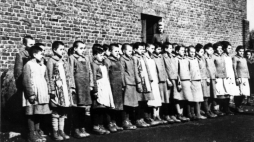 Więźniarki na apelu, stojące na tle ceglanego baraku. Za nimi nadzorczyni Eugenia Pohl – obóz przy ul. Przemysłowej w Łodzi, wiosna 1944 r. Źródło: IPN w Łodzi