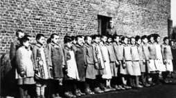 Więźniarki na apelu, stojące na tle ceglanego baraku. Za nimi nadzorczyni Eugenia Pohl - obóz przy ul. Przemysłowej w Łodzi, wiosna 1944 r. Źródło: IPN w Łodzi