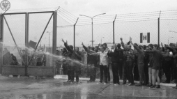 Akcja protestacyjna internowanych w ośrodku odosobnienia w Kwidzynie w dniu 14 sierpnia 1982 r. Polewani są wodą prawdopodobnie z węża strażackiego. Część z nich ma uniesione ręce z palcami w geście "V". Na drugim planie, wysoki płot za nim także stoją mężczyźni. Z lewej strony zamknięta brama. Źródło: Archiwum IPN