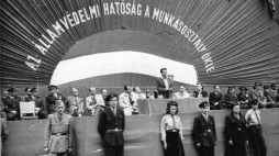 Węgierska Fabryka Wagonów i Maszyn (MÁVAG), 25 kwietnia 1949. Zdjęcie wykonano podczas przekazania flagi podarowanej przez pracowników zakładu Organowi Bezpieczeństwa Państwowego Ministerstwa Spraw Wewnętrznych (BM ÁVH). Cywile od lewej do prawej: Sándor Zöld, Ferenc Donáth, Mihály Farkas, János Kádár przy stole. Mówca István Hidas, Gábor Péter i matka flagi Mihály Farkas (Käthe Rüdiger)./Źródło: hu.wikipedia