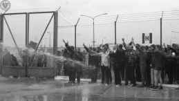 Akcja protestacyjna internowanych w ośrodku odosobnienia w Kwidzynie w dniu 14 sierpnia 1982 r. Polewani są wodą prawdopodobnie z węża strażackiego. Część z nich ma uniesione ręce z palcami w geście "V". Na drugim planie, wysoki płot za nim także stoją mężczyźni. Z lewej strony zamknięta brama. Źródło: Archiwum IPN