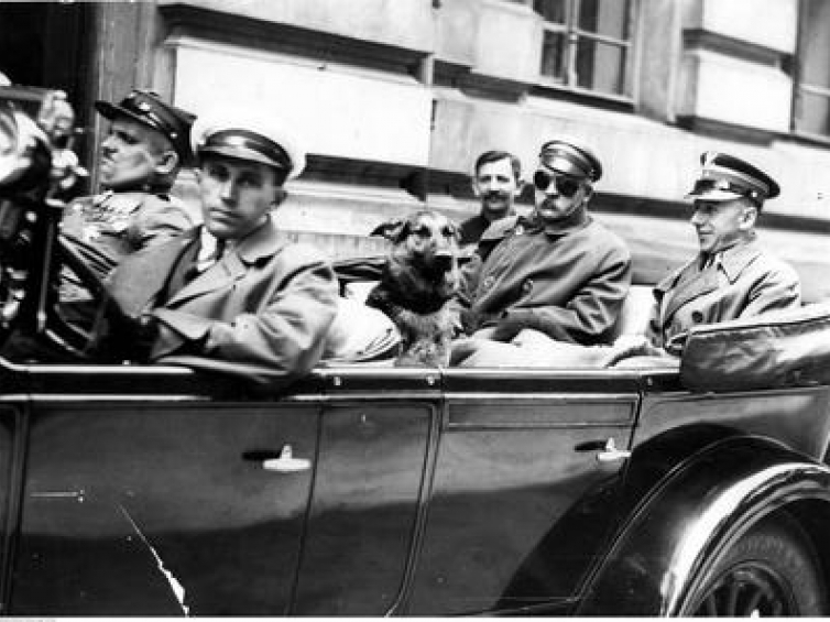 Na tylnym siedzeniu samochodu siedzą: Józef Piłsudski (po lewej stronie, w goglach), mjr Konrad Zembrzuski (z prawej) i pies "Pies". Samochód prowadzi szofer Zygmunt Malinowski, obok niego siedzi wachmistrz Walenty Wójcik. Źródło: NAC