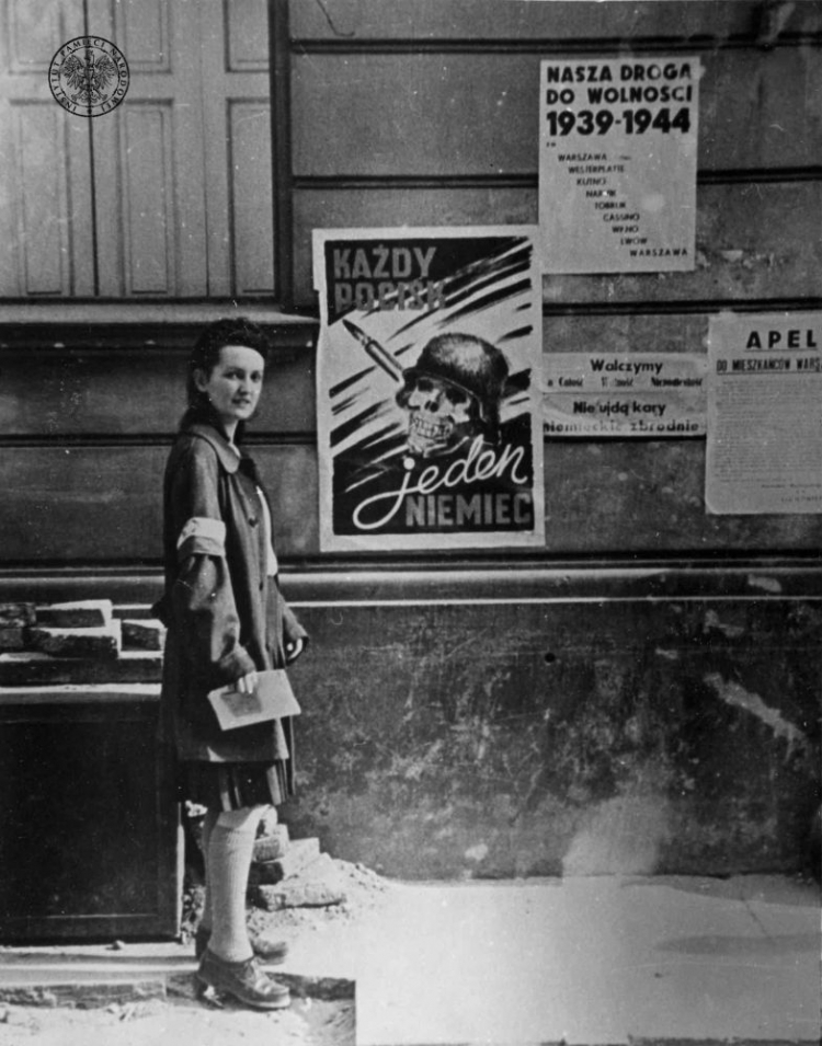 Łączniczka przy plakacie autorstwa Henryka Chmielewskiego „Każdy pocisk jeden Niemiec”. Warszawa, sierpień 1944 r. Autor: Eugeniusz Lokajski, fot. z zasobu IPN