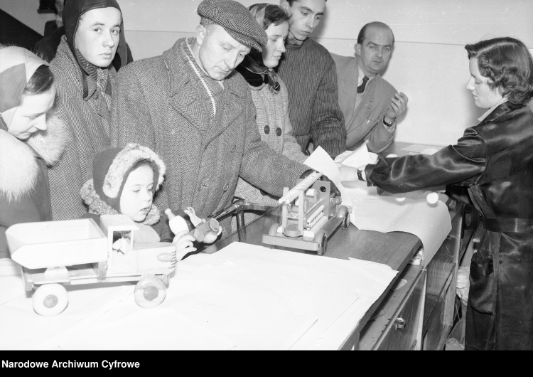 Sprzedawczyni pakująca zakupioną zabawkę. Na ladzie stoją: drewniany samochód strażacki oraz ciężarowy. Widoczni klienci oglądający zabawki, lata 1950 - 1960. /NAC