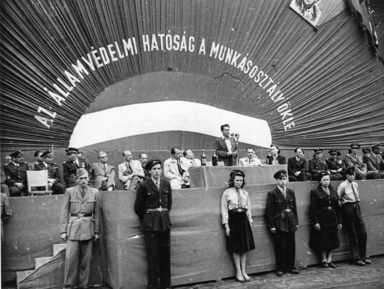 Węgierska Fabryka Wagonów i Maszyn (MÁVAG), 25 kwietnia 1949. Zdjęcie wykonano podczas przekazania flagi podarowanej przez pracowników zakładu Organowi Bezpieczeństwa Państwowego Ministerstwa Spraw Wewnętrznych (BM ÁVH). Cywile od lewej do prawej: Sándor Zöld, Ferenc Donáth, Mihály Farkas, János Kádár przy stole. Mówca István Hidas, Gábor Péter i matka flagi Mihály Farkas (Käthe Rüdiger)./Źródło: hu.wikipedia