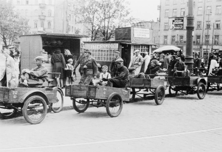 Reprodukcja zdjęcia Stefana Bałuka: Postój riksz na placu Żelaznej Bramy w Warszawie, lipiec 1944 r. Widok na południową część placu. W głębi widoczne kamienice u wylotu ulicy Granicznej: od prawej stoi budynek pl. Żelaznej Bramy 3 (róg Graniczna 17), potem (zasłonięty) wylot Granicznej i zburzony dom nr 5, po lewej budynek pl. Żelaznej Bramy 3. Fot. PAP/Reprodukcja